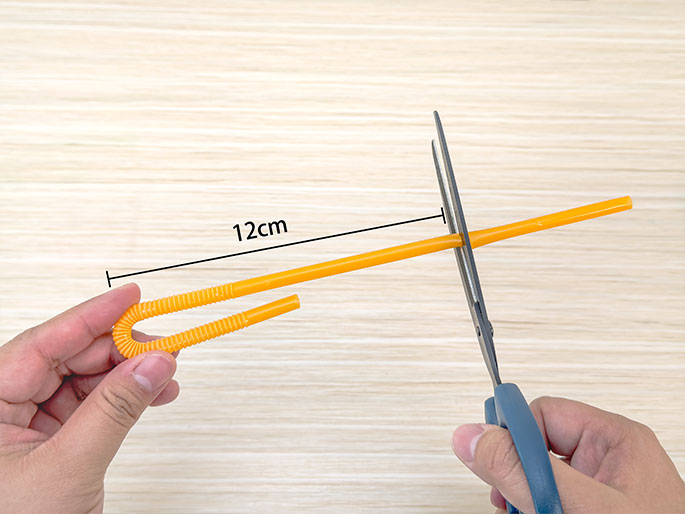 Two hands are shown on a light wood-grain background interacting with an orange drinking straw. One hand holds a shorter, bent section of the straw, while the other hand uses scissors to cut a longer, straight piece. A black line indicating 12cm spans the length of the longer straw segment, suggesting measurement and preparation for a craft or science activity.
