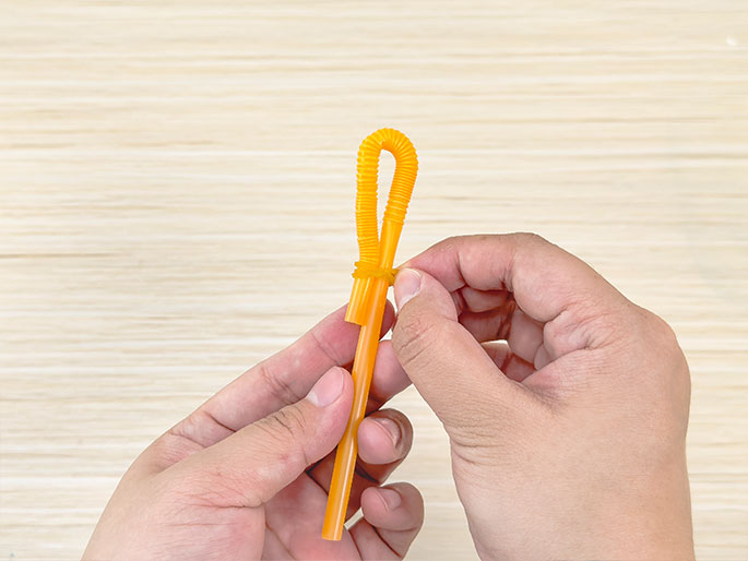 Two hands hold a flexible orange drinking straw against a light wood-grain background. The straw has been bent into a loop at the top and a small orange rubber band secures the base of the loop to the longer, straight portion of the straw. The hands appear to be adjusting or tightening the rubber band. The image suggests a craft project or the construction of a simple toy or tool.