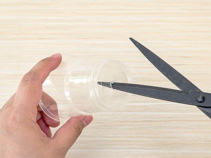 A hand holds the bottom portion of a clear plastic bottle against a light wood-grain background. Scissors are being used to make a small cut or notch in the center of the bottle's base. This implies the bottle is being prepared for a craft project or science experiment that requires modification.