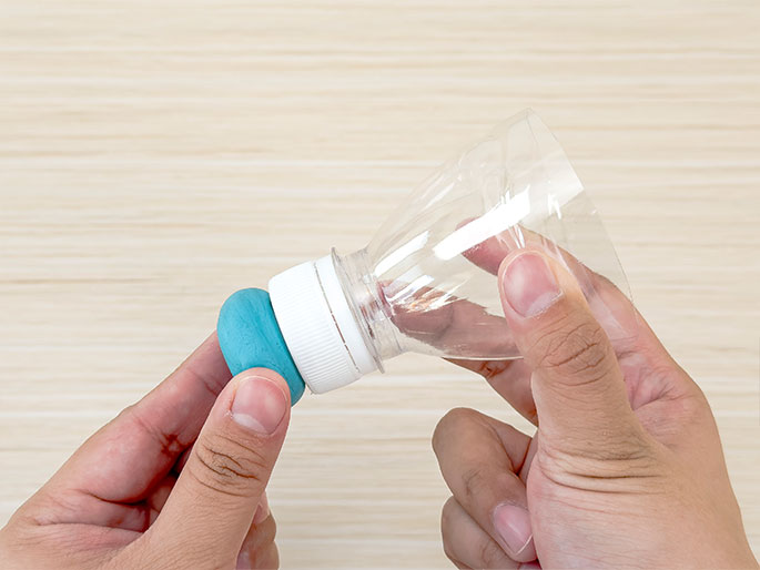 Two hands hold the top portion of a clear plastic bottle, including its white cap, against a light wood-grain background. A ball of turquoise putty or clay is affixed to the bottle cap. The image suggests the creation of a homemade device or toy using repurposed materials.