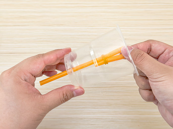 Two hands hold a clear plastic cup and an orange drinking straw against a light wood-grain background. A section of flexible orange straw, bent and secured with a small orange rubber band, is inserted into the cup. One end of the straight straw protrudes from the cup while the bent section remains inside, suggesting the construction of a simple pump, toy, or science experiment.