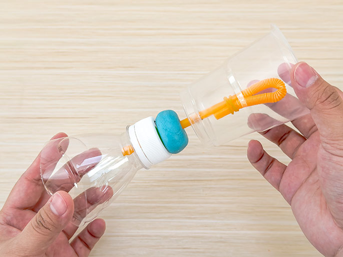 Two hands hold a clear plastic bottle and a clear plastic cup connected by an orange straw against a light wood-grain background. A ball of turquoise putty or clay is wrapped around the bottle's opening and the straw, creating an airtight seal. The flexible portion of the straw is inside the cup, and the setup resembles a homemade pump or air pressure experiment.