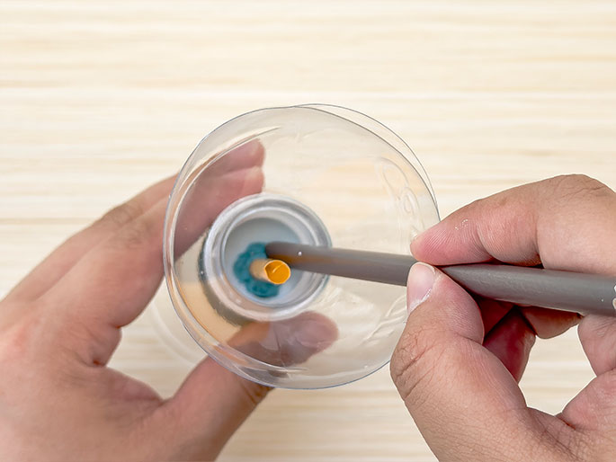 A hand holds the top, cut-off portion of a clear plastic bottle against a light wood-grain background. A white bottle cap, sealed with turquoise putty or clay, is visible inside the larger opening of the bottle. A gray pencil or similar cylindrical object is being inserted through the putty and into a small orange straw that is embedded within the putty. This suggests a science experiment or craft project involving airflow or pressure.