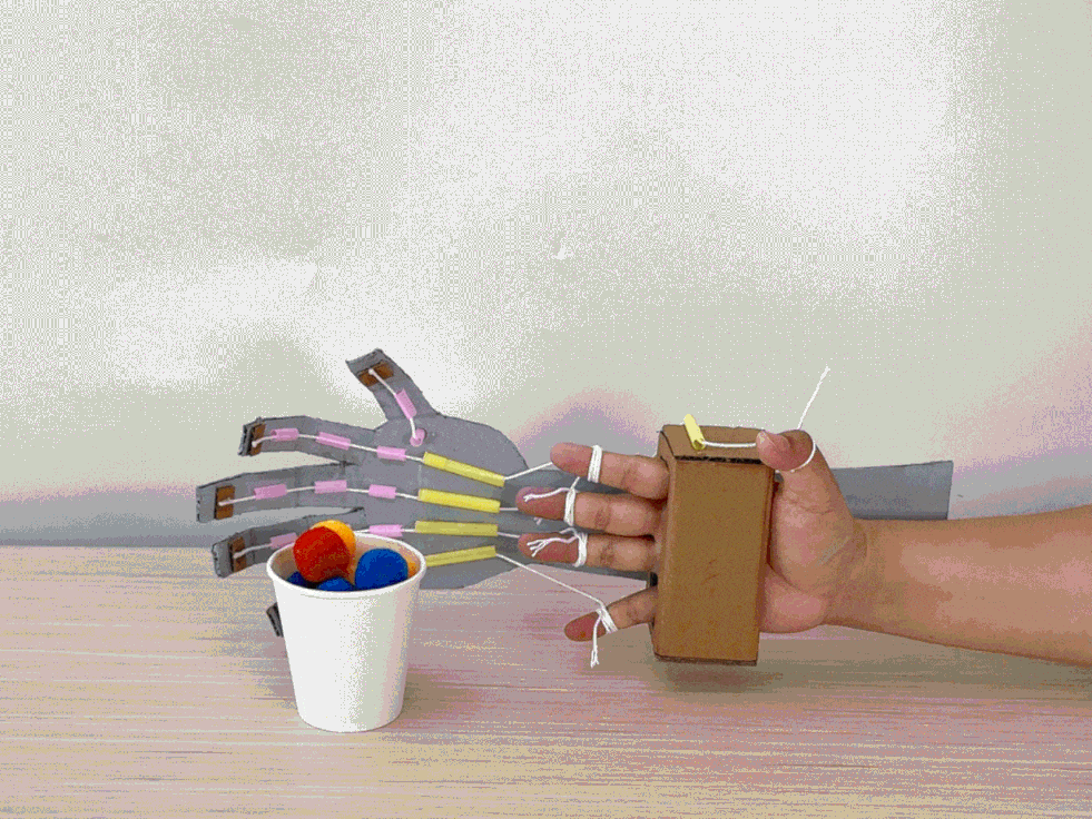 A child demonstrates a cardboard robotic hand. Strings connect the fingers of the cardboard hand to a small cardboard box worn on the child's hand. By moving their fingers inside the box, the child can control the cardboard hand to pick up and drop colorful pompoms.