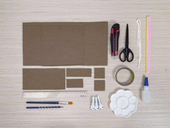 An assortment of craft supplies is arranged on a light wood-grain table, laid out in preparation for a project. The supplies include various sizes of cut cardboard pieces, a utility knife, scissors, string, colorful straws, a ruler, a paintbrush, a pencil, tubes of paint or glue, a flower-shaped paint palette, and a bottle of white glue. This image depicts the materials gathered for a crafting activity.