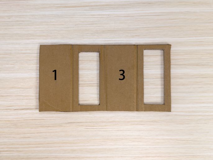 A piece of brown cardboard, divided into four sections and numbered 1 through 4, rests on a light wood-grain table. Rectangular holes have been cut out of sections 2 and 4. The remaining sections, 1 and 3, are solid. This image likely depicts a stage in a craft or DIY project involving cardboard construction.