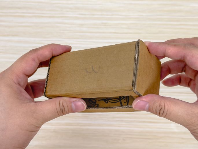 Two hands hold a small, rectangular box constructed from brown cardboard against a light wood-grain background. The box is open at one end, revealing a decorative pattern or design on the interior bottom. The hands gently support the box, showcasing its simple construction. This image likely illustrates a finished or nearly finished DIY cardboard craft project.