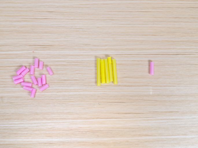 On a light wood-grain table, several small, colorful plastic tubes are arranged in three groups. A loose pile of pink tubes is on the left, a neat row of yellow tubes is in the center, and a single pink tube is on the right. These tubes appear to be components for a craft or educational activity.