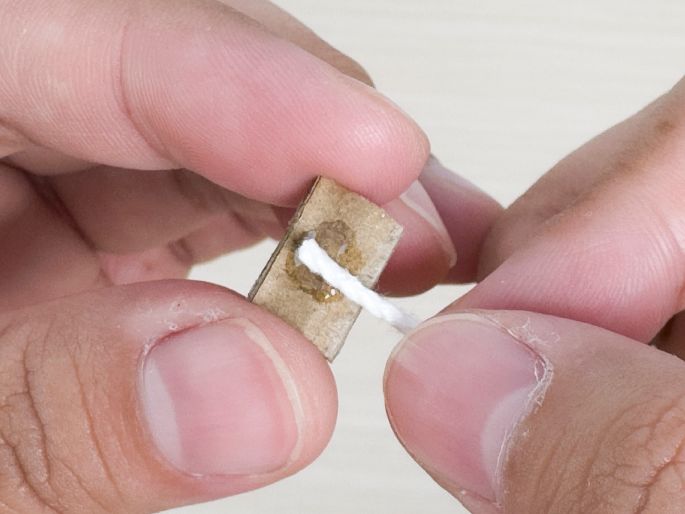 Close-up of two hands holding a small piece of cardboard. A length of white string or twine is being glued or attached to the cardboard using a clear adhesive. The focus is on the process of securing the string to the cardboard.