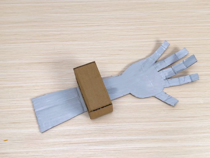 A simple robotic arm crafted from cardboard lies on a light wood-grain surface. The arm is painted silver and has segmented fingers, giving it a robotic appearance. A small, unpainted cardboard box is attached to the arm, serving as a control panel or joint. This image likely depicts a children's craft project or a simple robotics model.