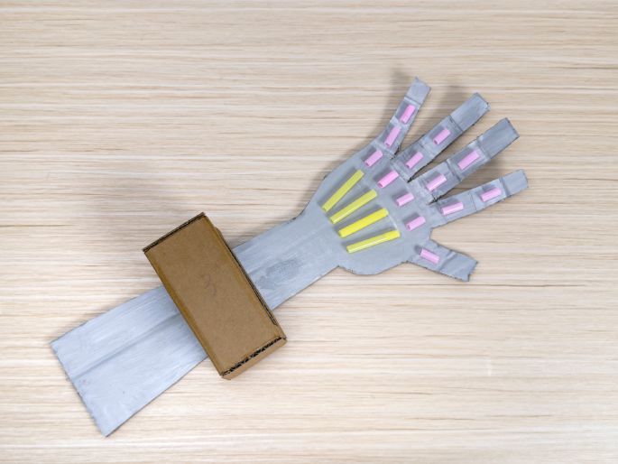A cardboard robotic arm, painted silver, rests on a light wood-grain table. Pink and yellow plastic tubes are attached to the hand section, representing finger joints or mechanical components. A small, plain cardboard box is affixed to the arm, likely symbolizing a control unit. This image showcases a completed DIY robotic hand craft project, likely made by a child.