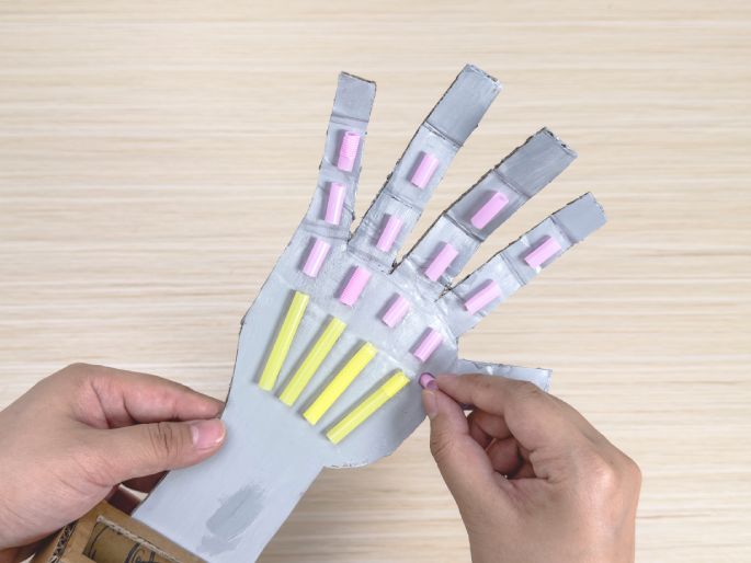 Two hands are working on a cardboard robotic hand against a light wood-grain background. One hand holds the silver-painted cardboard cutout of the robotic hand, while the other hand carefully places small pink tubes onto the hand shape. Yellow tubes are already affixed to the palm area. This image captures the process of assembling a children's craft project.