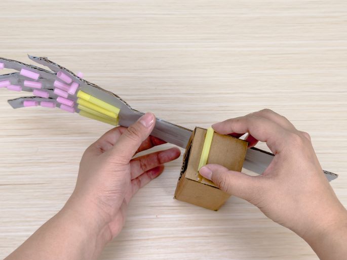 Two hands are assembling a cardboard robotic arm on a light wood-grain surface. One hand holds the silver-painted cardboard arm, which has pink and yellow tubes attached to the hand section. The other hand is attaching a yellow tube to a small, plain cardboard box, which will likely act as a joint for the arm. This image shows the construction phase of a DIY robotic arm craft project.