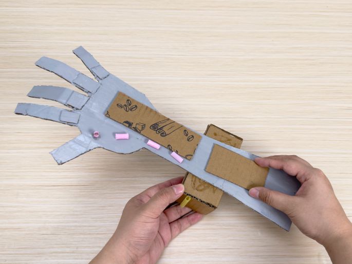 Two hands are assembling a cardboard robotic arm on a light wood-grain table. The arm is partially painted silver, with the hand section more fully painted than the forearm. Small pink tubes are attached to the hand, and a piece of unpainted cardboard with printed designs is affixed to the forearm. A small cardboard box acts as a joint, connecting a yellow straw to another part of the arm. This image captures the construction process of a DIY cardboard robotic arm, likely a children's craft project.