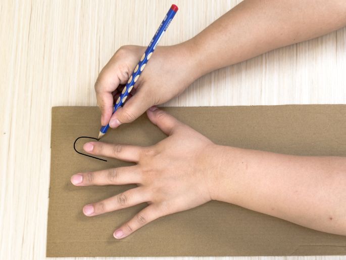 Two hands are positioned on a piece of brown cardboard on a light wood-grain table. One hand holds a blue pencil and is tracing the outline of a finger from the other hand onto the cardboard. This image shows the process of creating a template or pattern by tracing a hand or finger shape onto cardboard.
