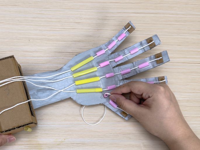 A partially constructed cardboard robotic hand is being worked on against a light wood-grain background. The hand is painted silver, with pink tubes attached to the fingers and yellow tubes along the palm. White strings are threaded through the hand and connect to a simple cardboard box that serves as the wrist. A hand is seen in the image, threading a string through one of the pink tubes. This image captures the process of creating a functional cardboard robotic hand, likely a science or engineering project.