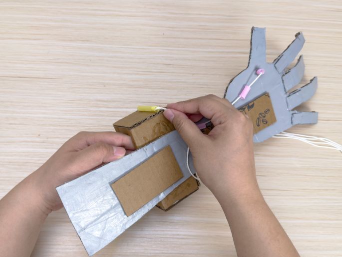Two hands are in the process of assembling a cardboard robotic arm on a light wood-grain surface. The arm is made from plain brown cardboard and sections painted silver. White strings are being threaded through the wrist area, which is a small box-like structure. The hand portion, visible in the background, has pink and yellow tubes attached to the fingers and palm, also connected by strings. This image shows the construction of a DIY robotic hand, likely a science or engineering project.