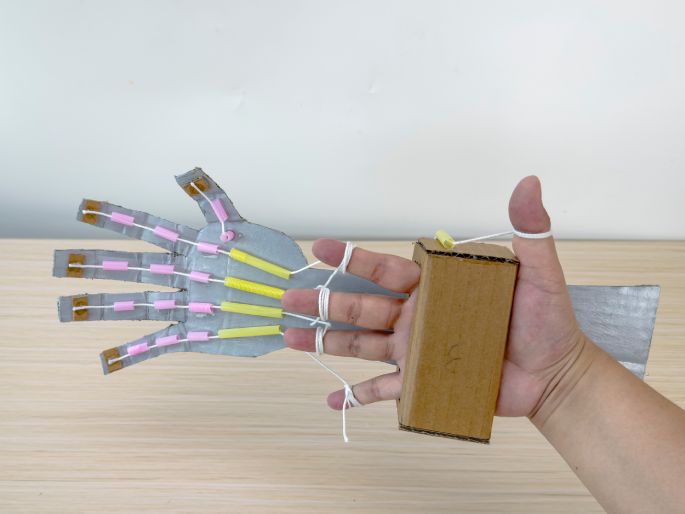 A hand is demonstrating a completed cardboard robotic hand against a plain, light-colored background. The robotic hand is made of silver-painted cardboard with pink and yellow straws attached to the fingers and palm. White strings connect the fingers to a small cardboard box worn on the back of the hand, acting as the control mechanism. The hand is positioned to show how pulling the strings manipulates the cardboard fingers. This image showcases a finished DIY robotic hand project, likely created for educational or demonstration purposes.