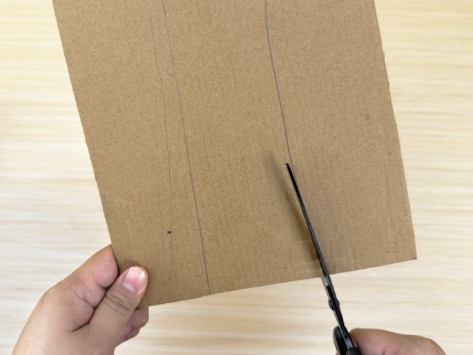 Two hands are shown working with a piece of brown cardboard on a light wood-grain table. One hand holds the cardboard, which has several lines drawn on it. The other hand holds a pair of black scissors and is in the process of cutting along one of the lines. This image illustrates the step of cutting cardboard along pre-drawn lines, likely for a craft or construction project.