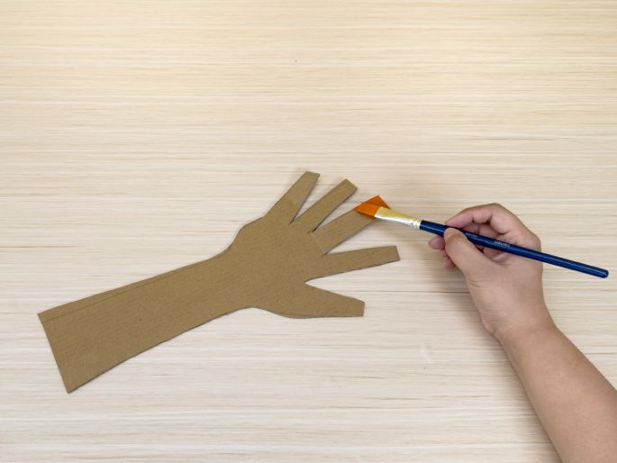 A hand-shaped piece of cardboard lies on a light wood-grain table. Another hand holds a paintbrush with orange paint and is applying the paint to the 'fingers' of the cardboard cutout. This image shows the process of painting a cardboard hand, likely for a craft project.