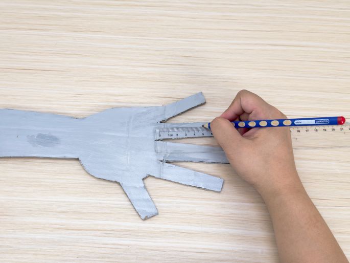 A silver-painted cardboard hand cutout rests on a light wood-grain table. A hand holding a pencil is using a clear ruler to draw straight lines across the fingers of the cardboard hand. This image captures the step of marking precise lines for cutting or folding on a craft project.