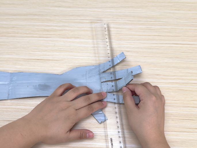 Two hands manipulate a silver-painted cardboard hand cutout on a light wood-grain table. The 'fingers' of the cardboard hand have been partially cut and are being gently bent. A clear ruler is positioned alongside the cuts, likely used as a guide for the bending or cutting process. This image shows the stage of shaping the cardboard hand for a craft project.