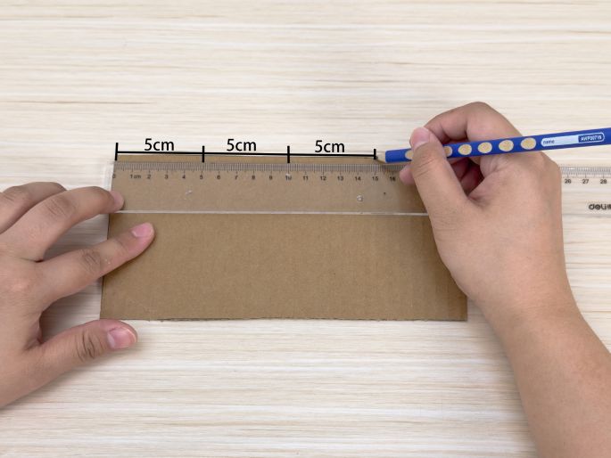 On a light wood-grain table, a piece of brown cardboard is being marked with a pencil. A hand uses a clear ruler to guide the pencil, marking three 5cm increments along the cardboard's edge. This image shows precise measurement and marking for cutting or folding cardboard, likely for a craft or DIY project.