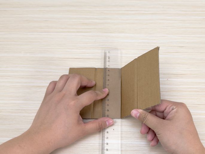 Two hands are folding a piece of brown cardboard along a scored line on a light wood-grain table. A clear ruler is placed along the fold, assisting in creating a crisp, straight edge. This image demonstrates the process of folding cardboard precisely, likely for constructing a small box or other cardboard structure.
