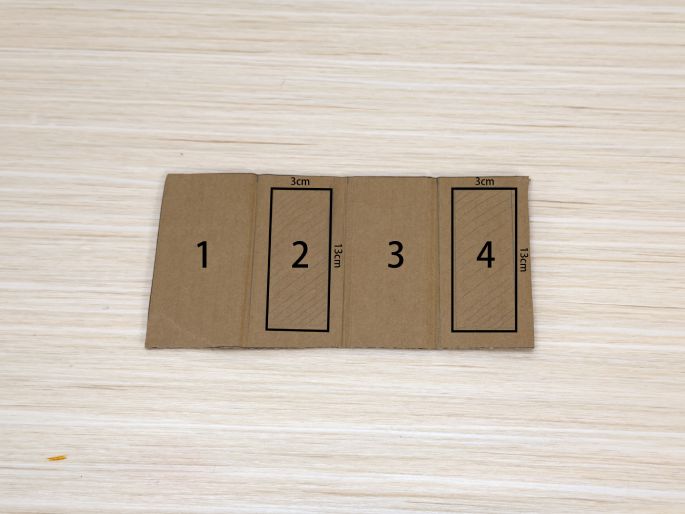A piece of brown cardboard lies flat on a light wood-grain table. It's divided into four sections, numbered 1 through 4. Sections 2 and 4 have rectangular outlines drawn within them, measuring 3cm wide and 13cm long, as indicated by labels. This image appears to show a template or plan for cutting or folding the cardboard.