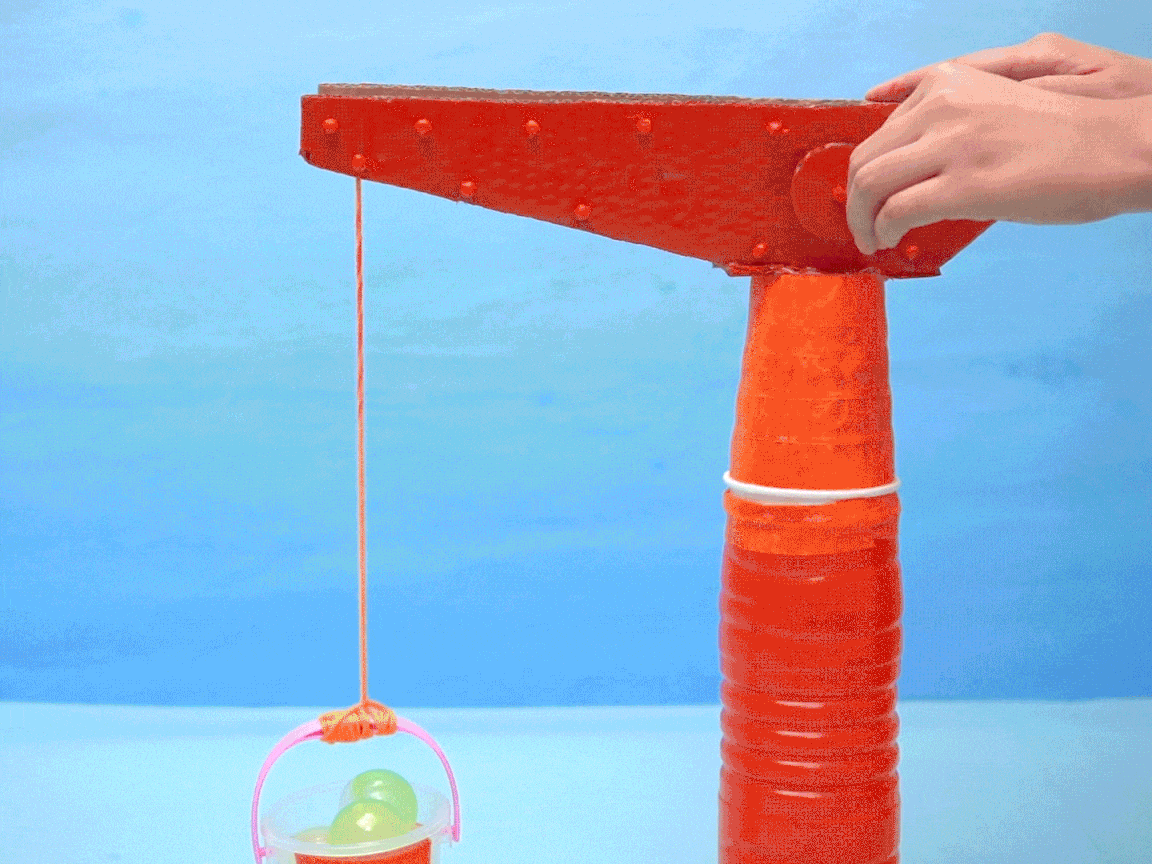 A small, homemade crane, constructed from cardboard and plastic cups, is operated by hand to lift a miniature bucket containing water beads or similar objects.