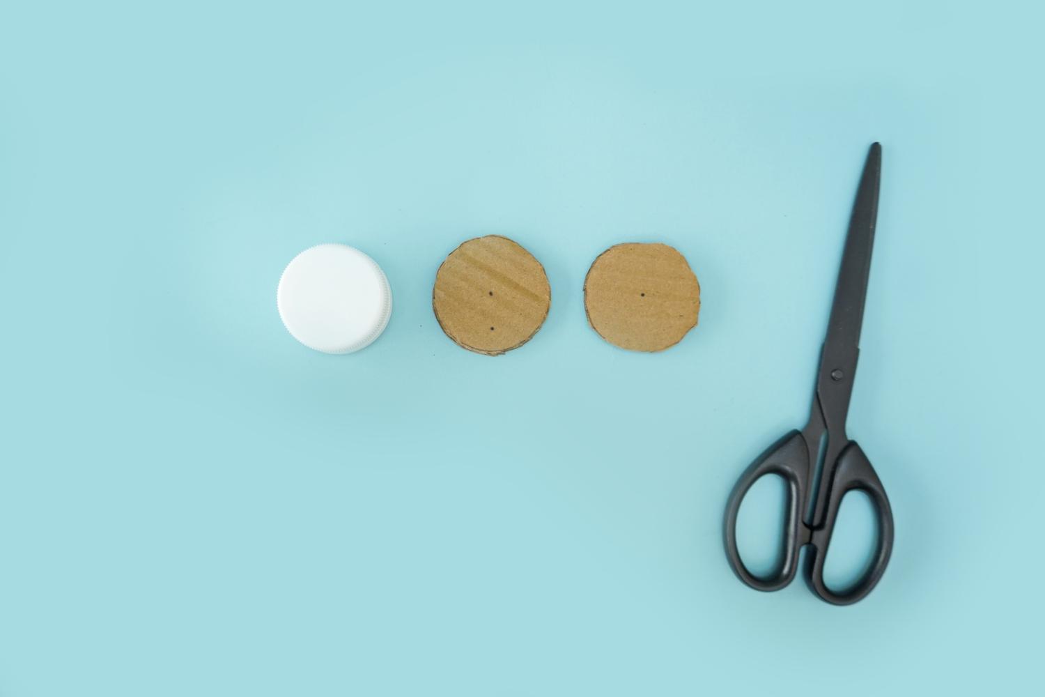 A white bottle top and two circles of cardboard are arranged on a pale blue surface next to a pair of black scissors.