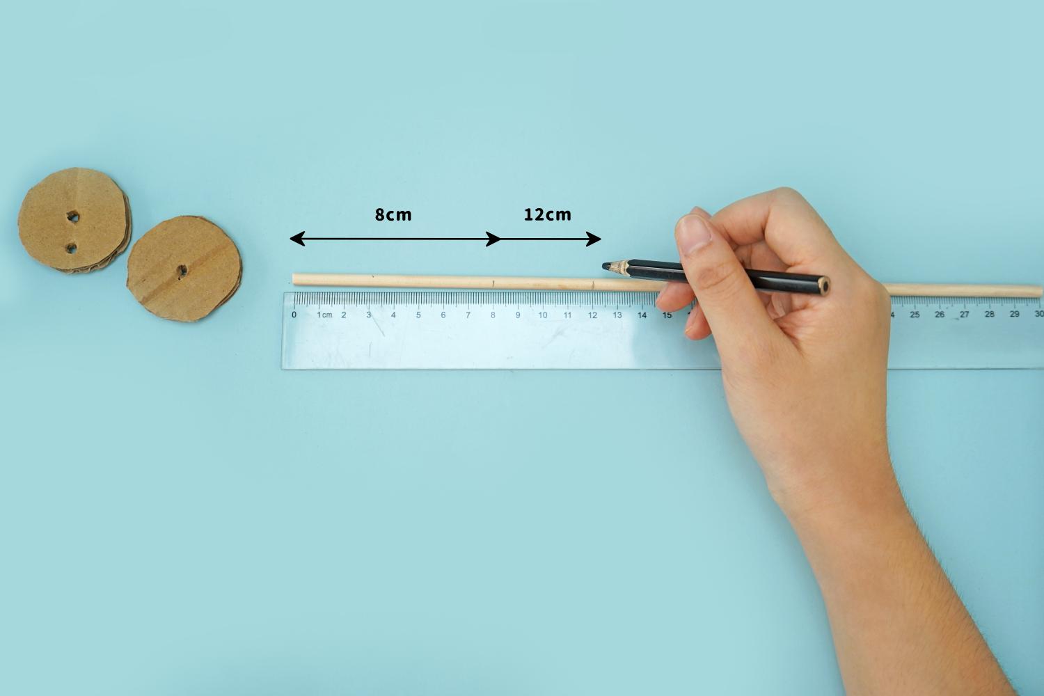 A hand uses a pencil to mark a 12cm length on a wooden stick, next to a ruler and two small cardboard circles, on a pale blue surface.