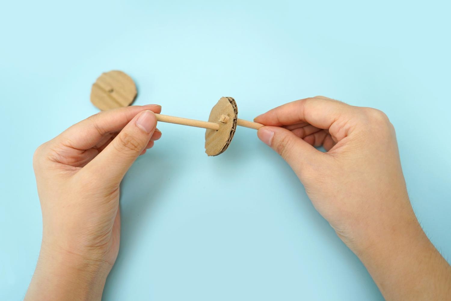 Two hands carefully attach a small wooden stick through the centre of a cardboard circle on a pale blue surface.