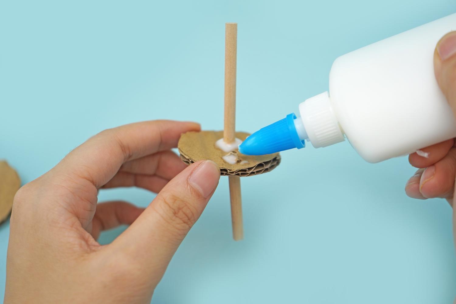 A hand applies glue to a cardboard circle attached to a wooden stick on a pale blue surface.