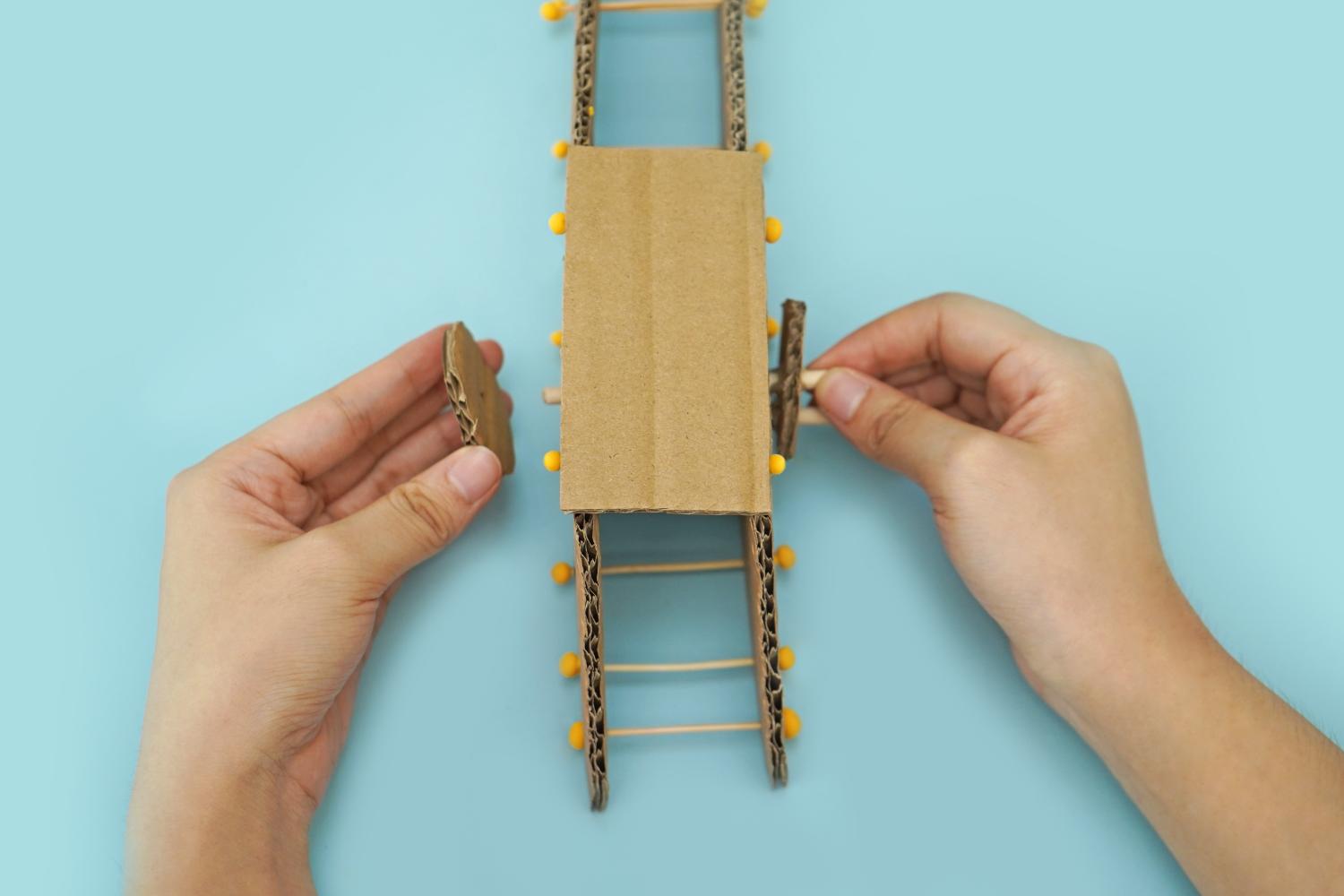 Two hands attach a small cardboard piece to a larger cardboard structure, which includes toothpicks and yellow modelling clay balls, on a pale blue background.