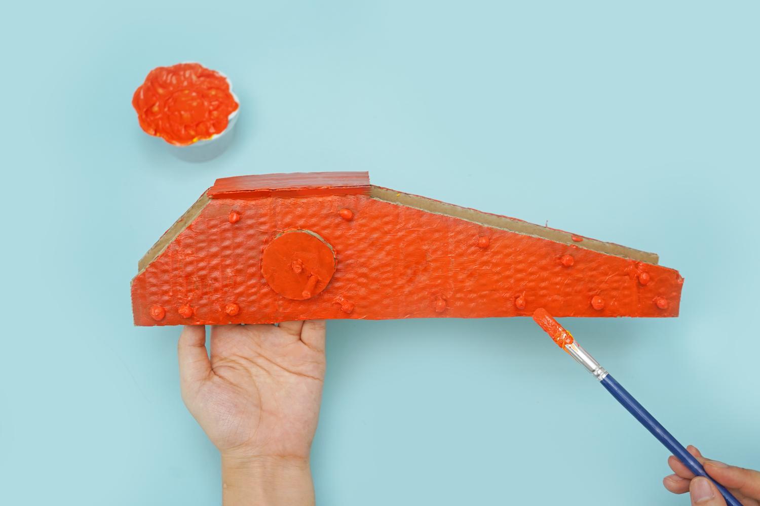 A hand holds a cardboard crane arm, already partially painted orange, whilst another hand uses a brush to add more orange paint, against a pale blue background.