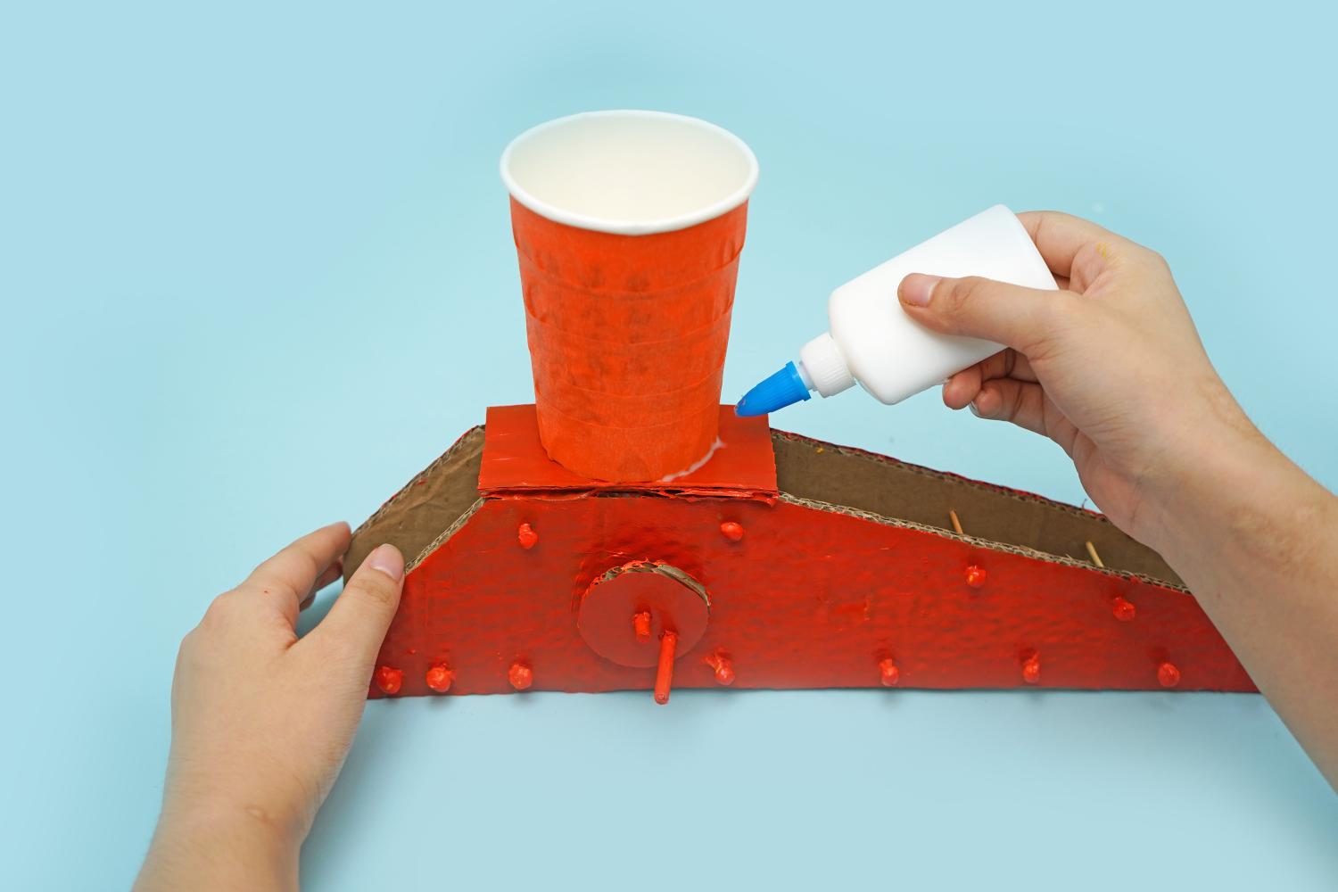 A hand applies glue to secure an orange paper cup to a painted red cardboard crane structure on a pale blue surface.