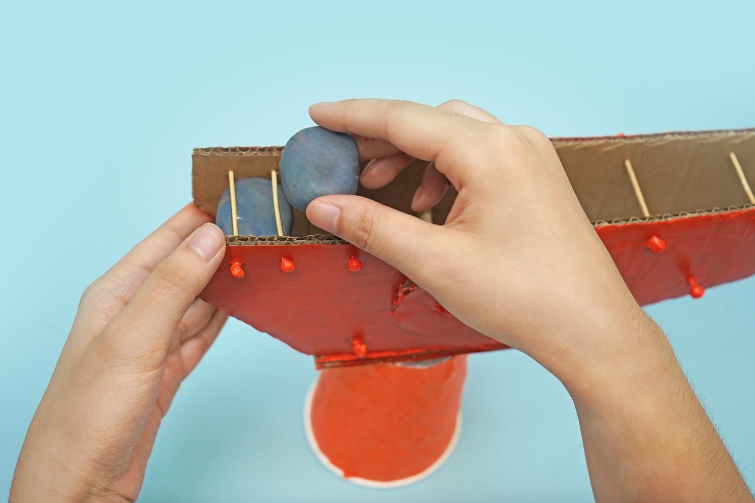 Two hands place small balls of grey modelling clay into a section of a red painted cardboard crane structure, supported by toothpicks, above an orange cup.