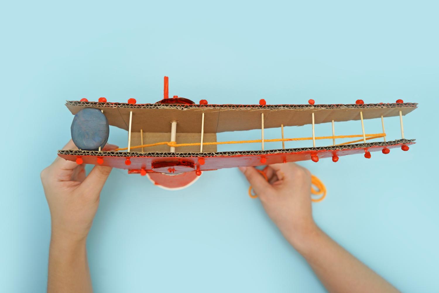 Two hands add a length of yellow elastic to a red painted cardboard crane model, which includes toothpicks and a grey modelling clay weight, on a pale blue surface.
