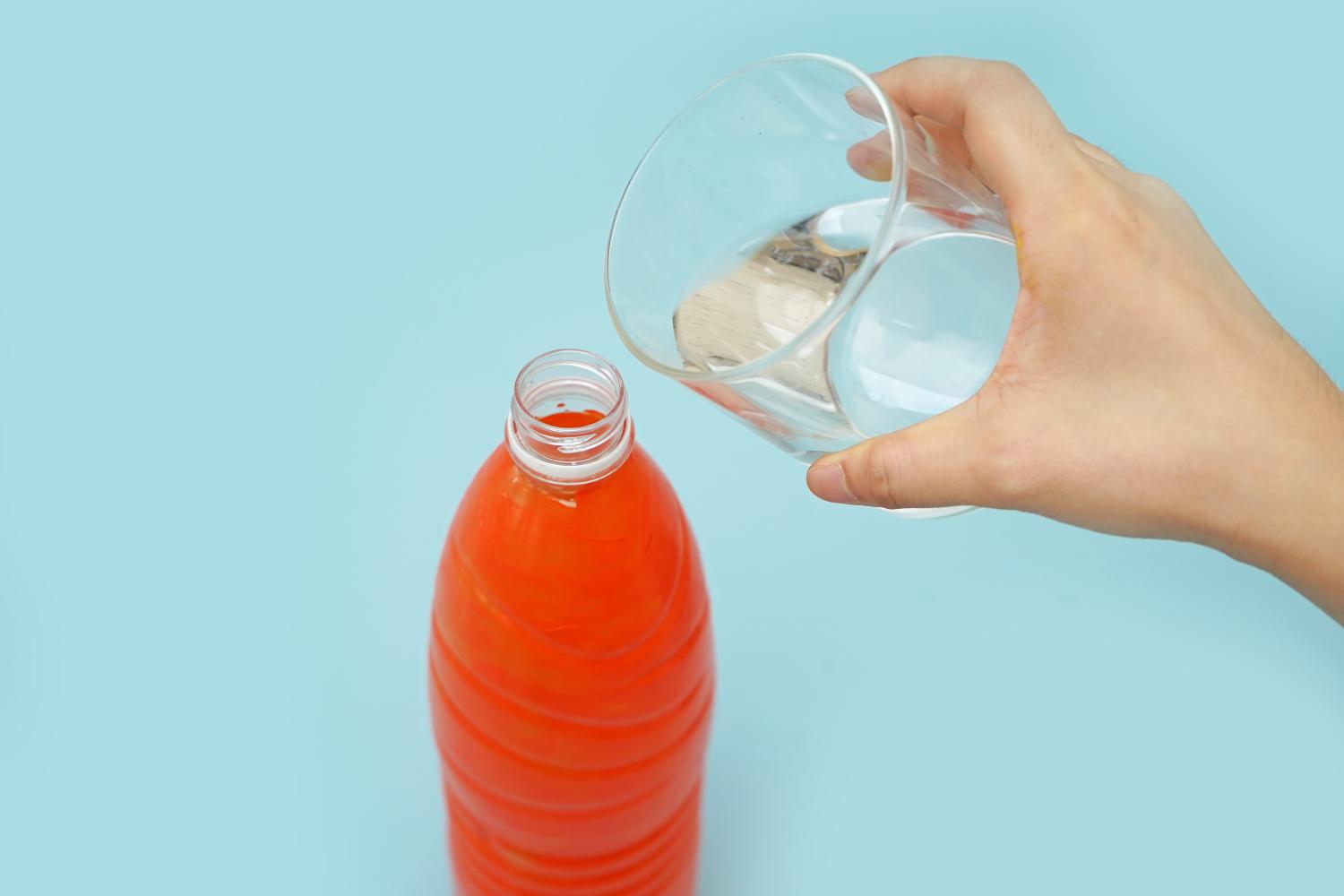 A hand pours water from a glass into an orange plastic bottle on a pale blue background.