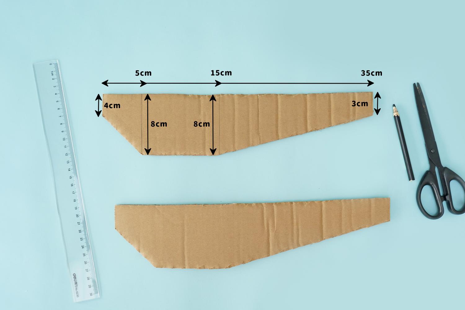 Two pieces of cardboard, marked with measurements in centimetres, are shown alongside a ruler, pencil, and scissors on a pale blue surface, ready for cutting.