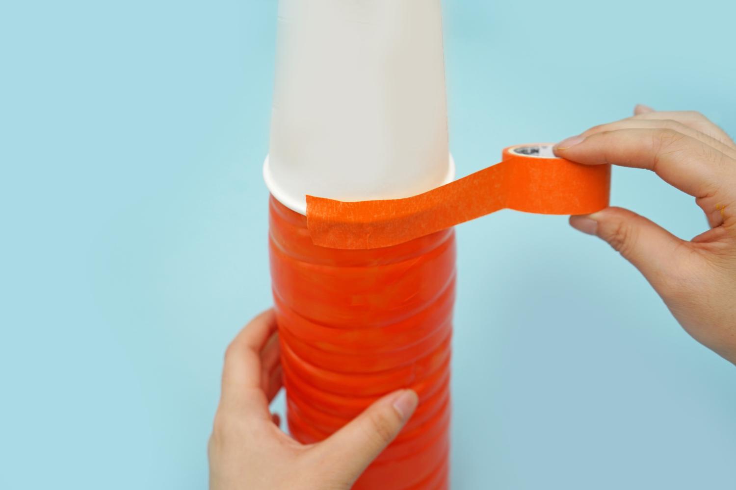 A hand uses orange tape to secure a white paper cup to an orange plastic bottle on a pale blue surface.