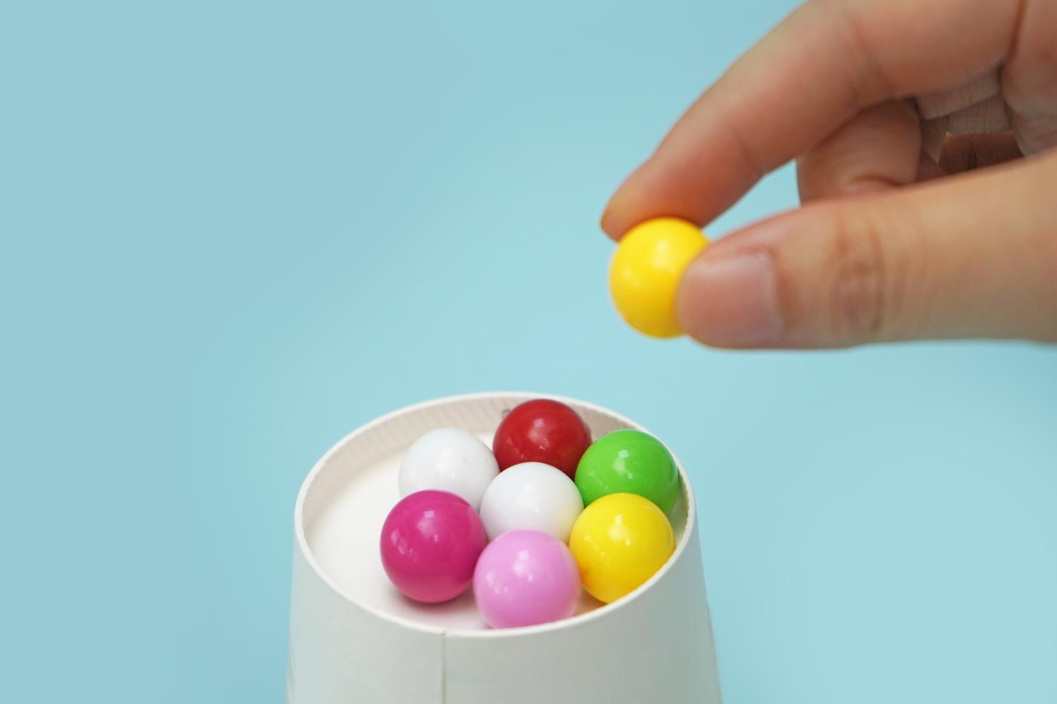 A hand adds a yellow sweet to a group of similarly-sized sweets in various colours, arranged in a white cup on a pale blue background.