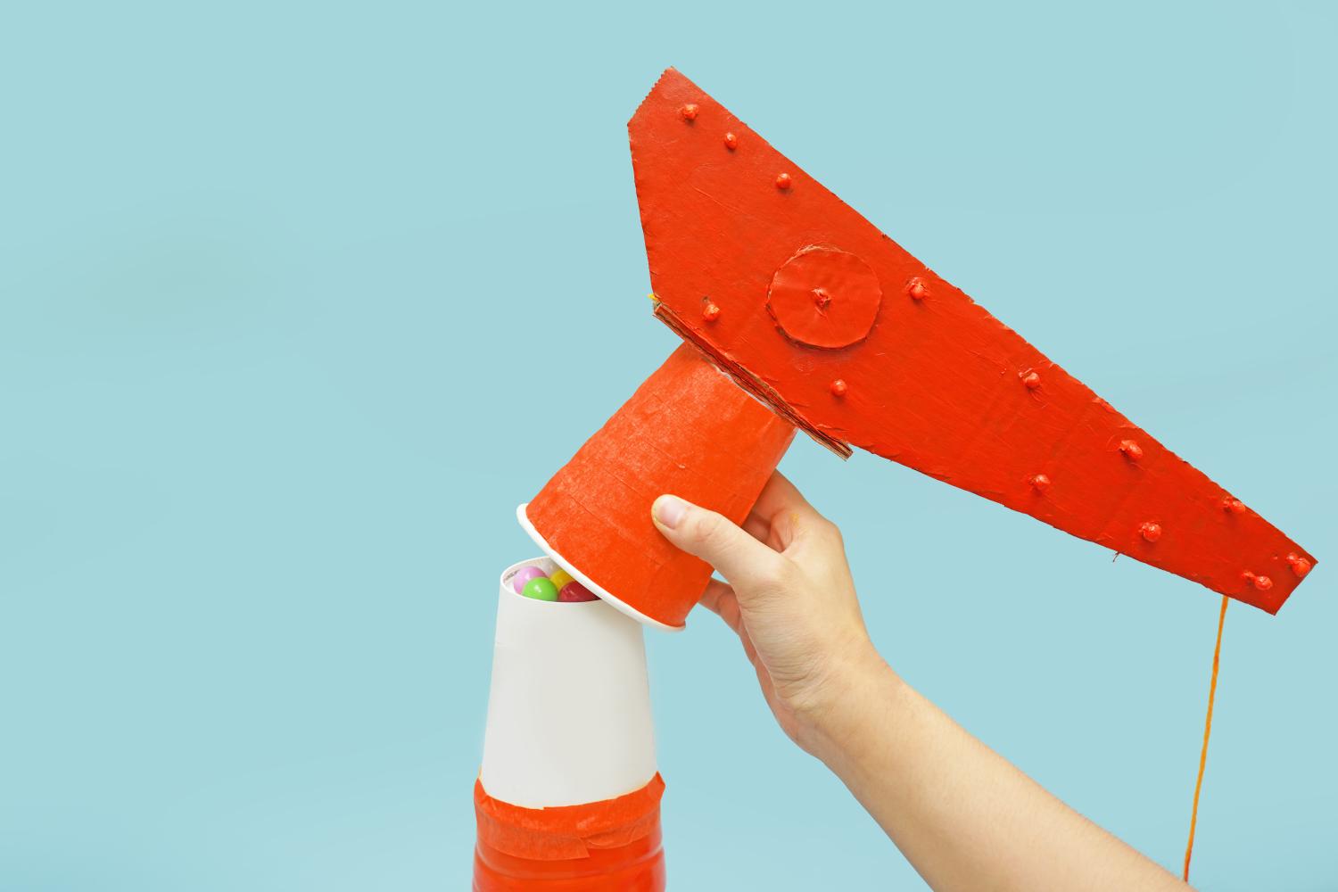A hand lifts a painted orange paper cup from a cardboard crane mechanism, revealing sweets inside a smaller cup below, against a pale blue background.
