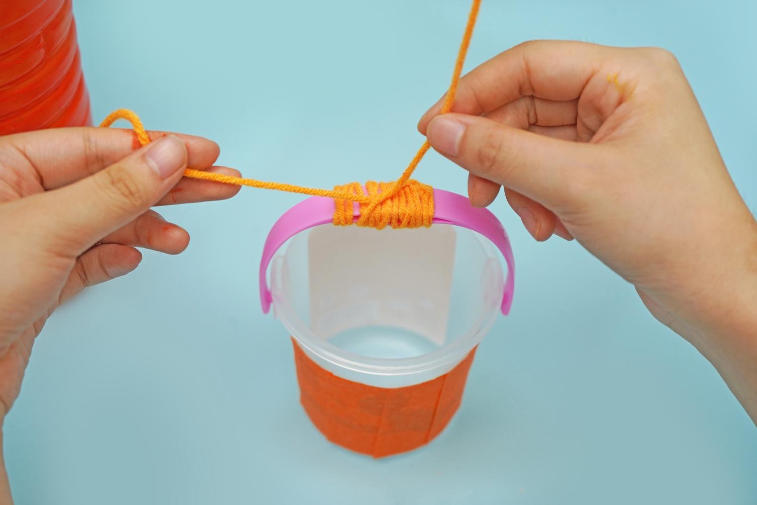 Two hands wrap orange yarn around the handle of a small plastic bucket on a pale blue surface.