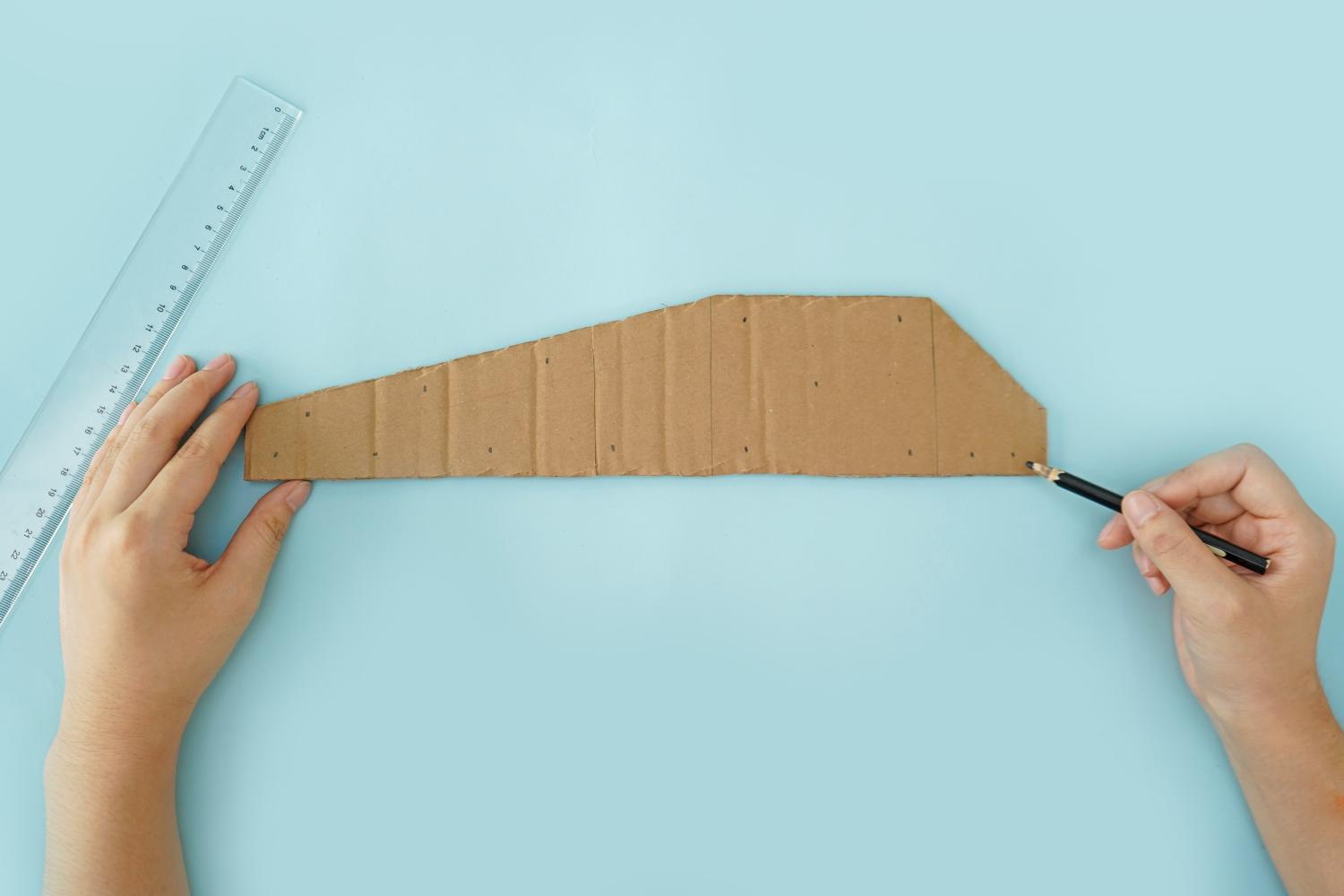 Two hands use a pencil and ruler to mark points on a piece of cardboard on a pale blue surface, preparing it for cutting.
