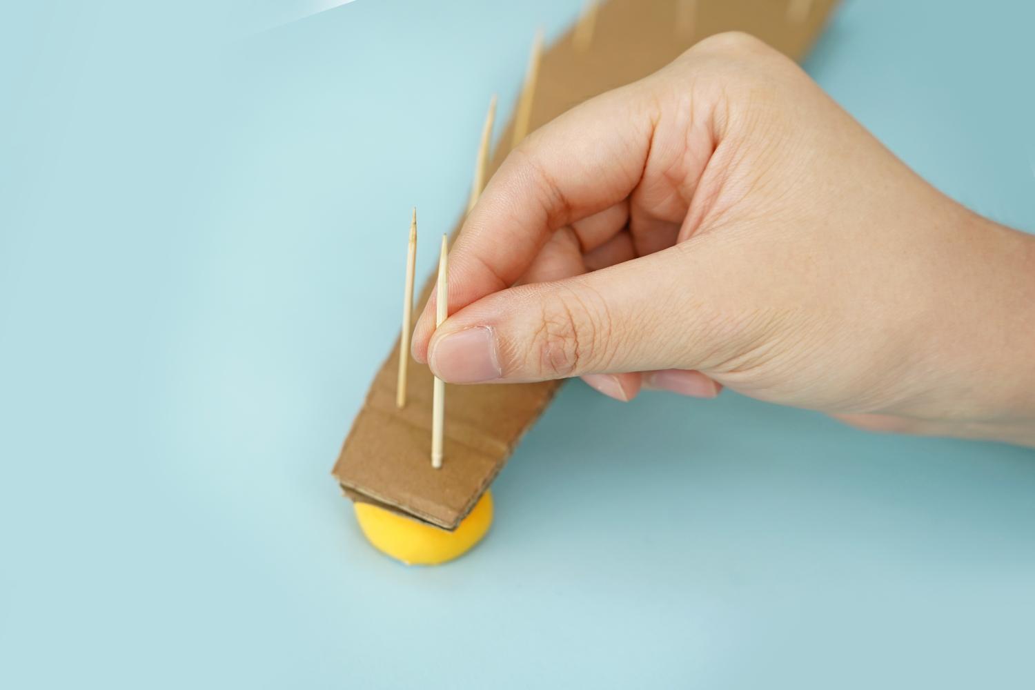 A hand carefully inserts toothpicks into a piece of cardboard resting on a yellow blob of modelling clay on a pale blue surface.