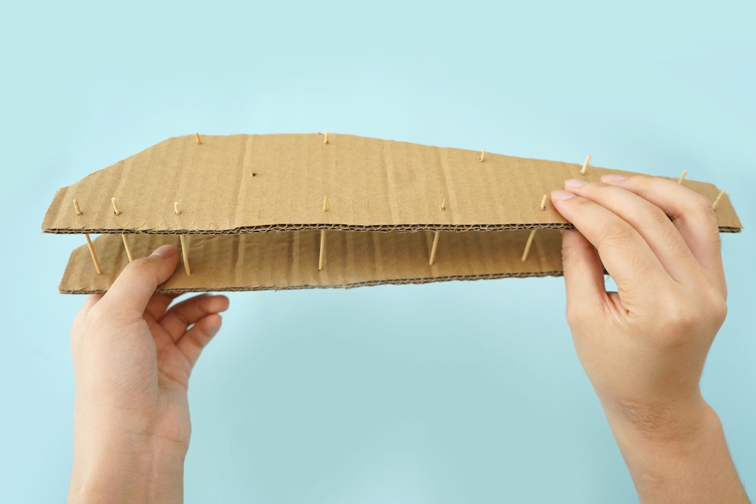 Two hands hold a section of cardboard structure, with toothpicks inserted vertically between two layers, against a pale blue background.
