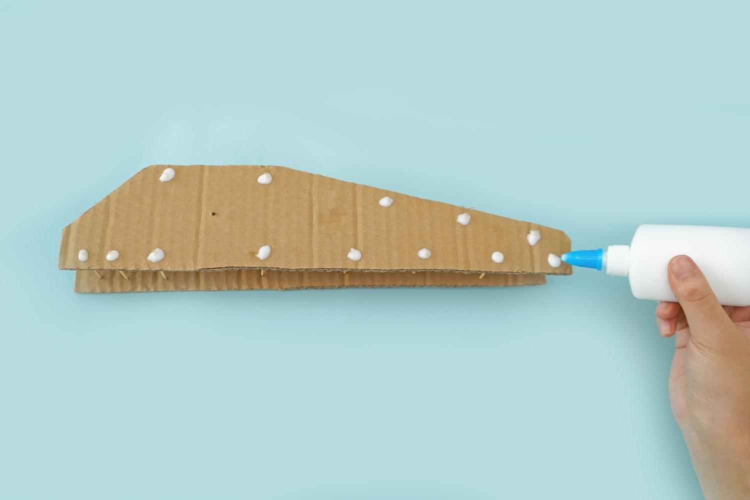 A hand applies white glue to a cardboard structure with toothpicks inserted, on a pale blue surface.
