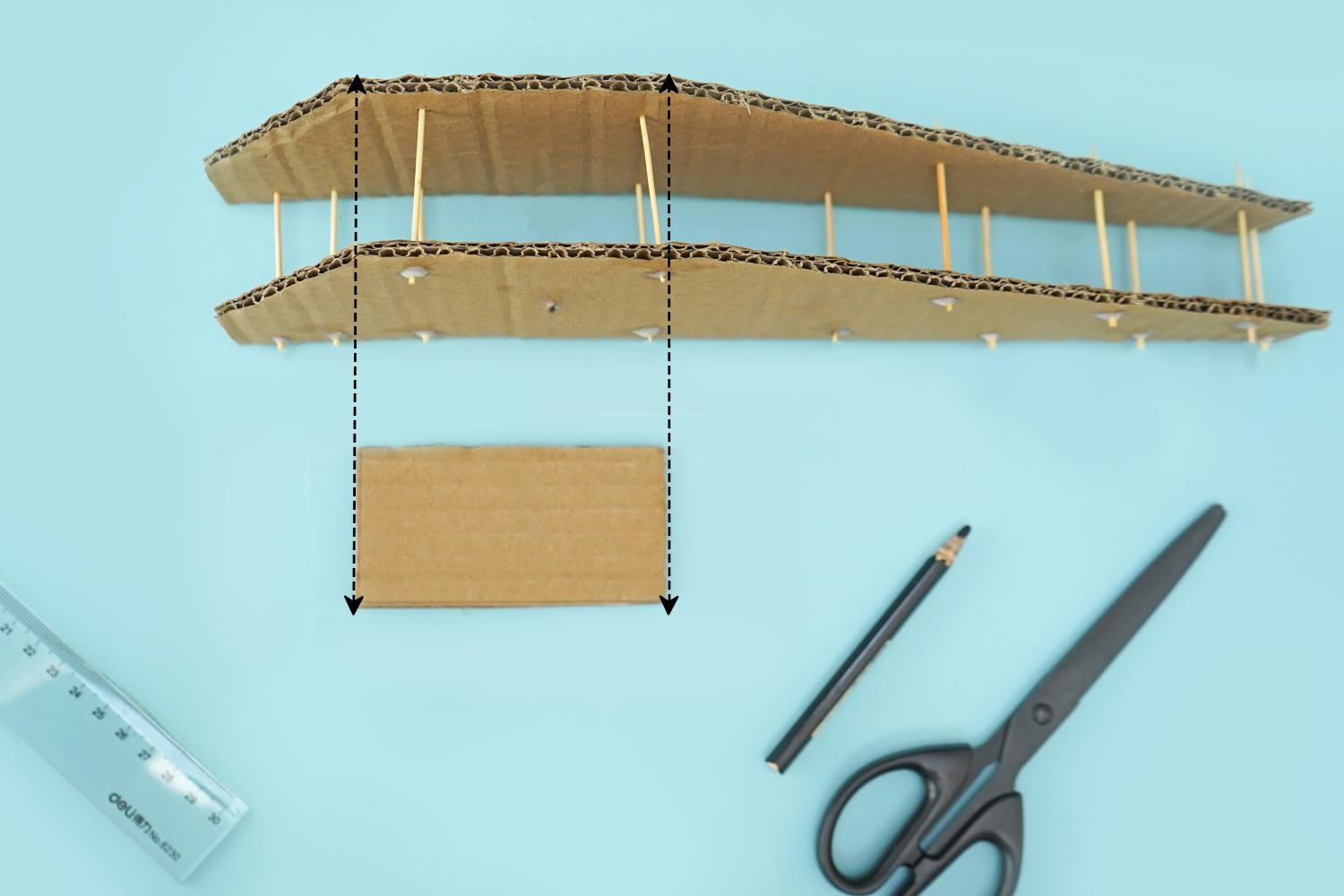 A piece of cardboard, with toothpicks glued between two layers, is shown next to a smaller piece of cardboard and a ruler, pencil and scissors on a pale blue surface.